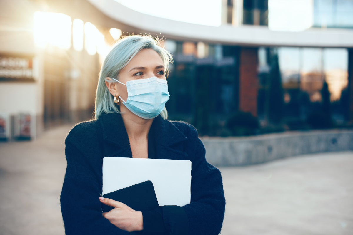 Careful caucasian businesswoman holding a laptop and tablet while wearing a protective mask